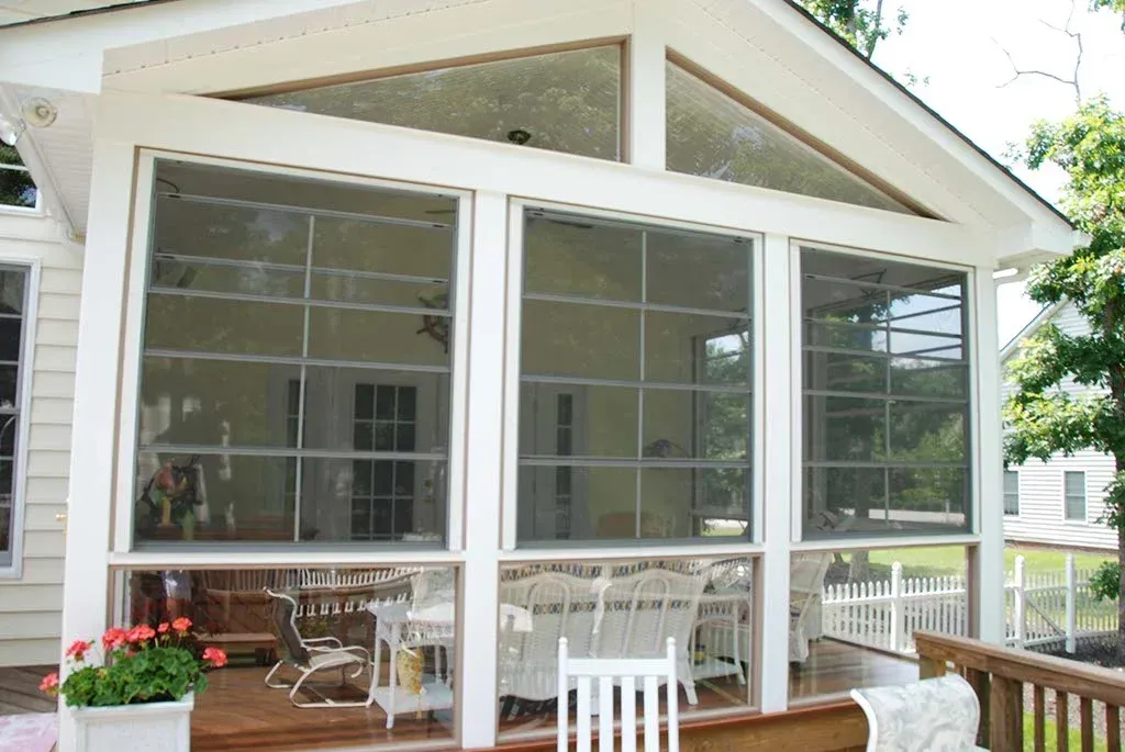 Screened porch with large windows, white trim, and a wooden deck. A white picket fence is visible in the background.