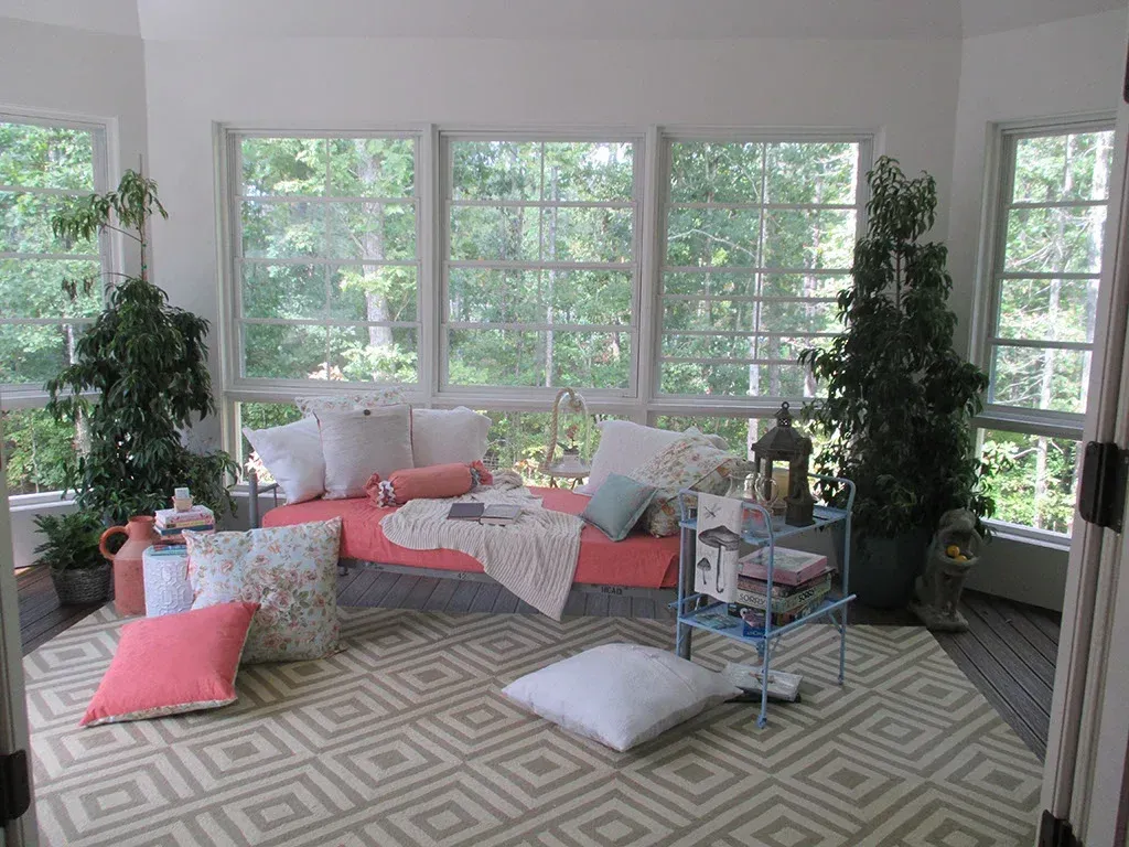 Sunroom with coral daybed, white pillows, patterned rug, and large windows overlooking trees.
