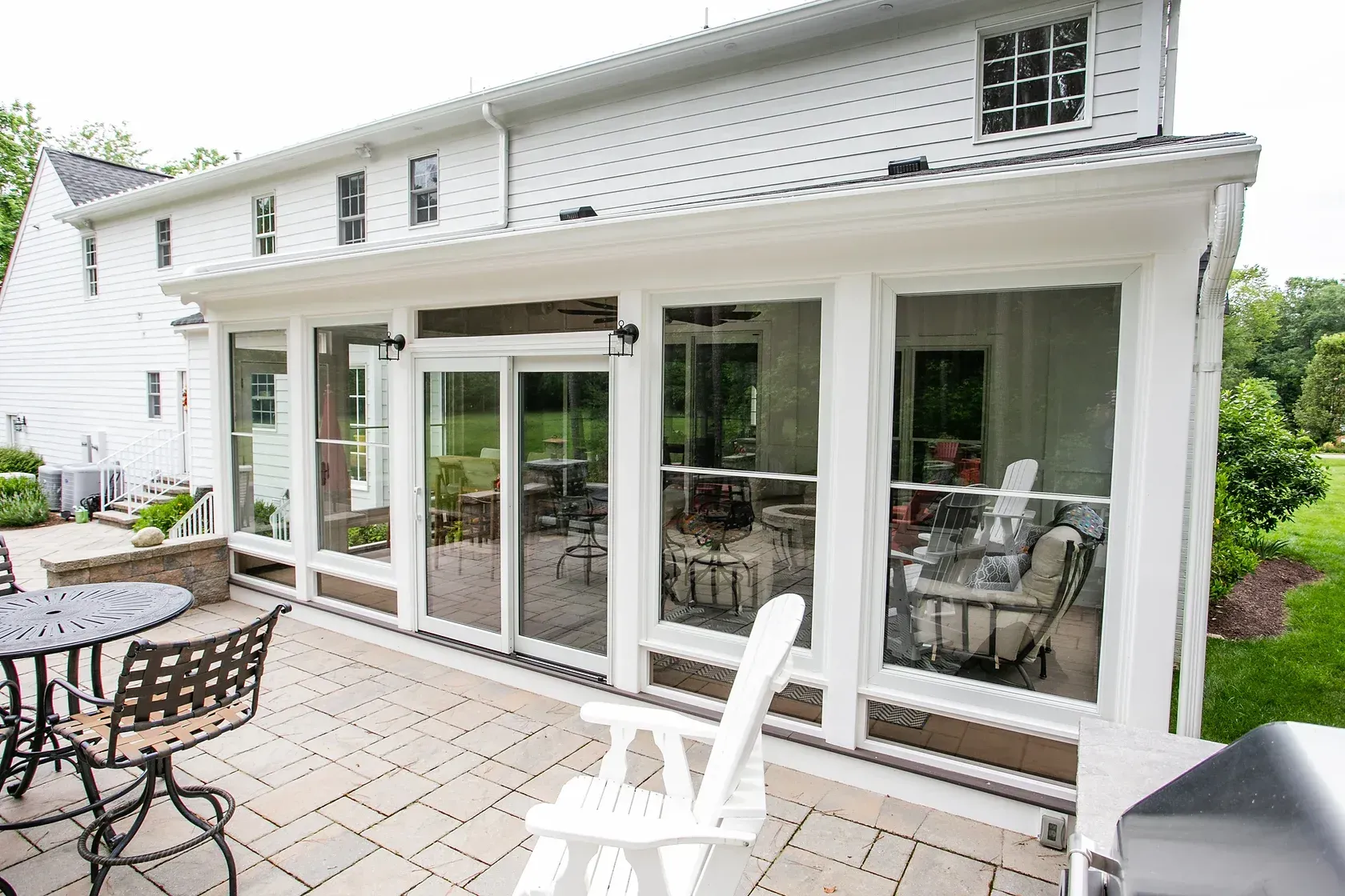 White-framed sunroom with glass walls attached to a white house, on a stone patio with outdoor furniture.