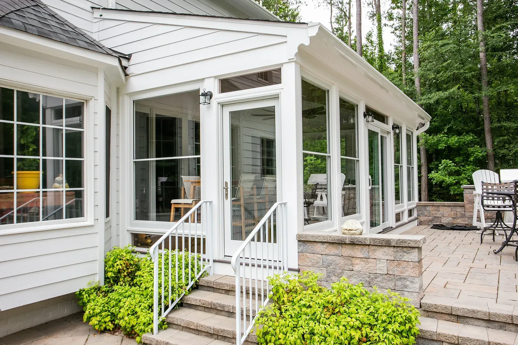 White-framed sunroom with glass walls, doors, and a stone facade. Steps lead to a patio with outdoor furniture.