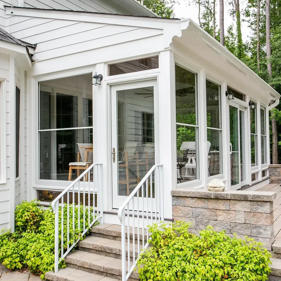 White sunroom with glass windows and door, stone patio, white railing and steps. Green bushes surround.