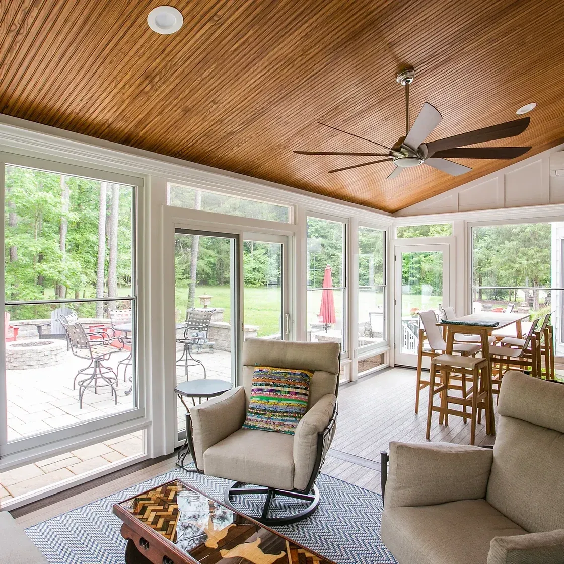 Sunroom with large windows, plush seating, and wooden ceiling, overlooking a patio and green backyard.
