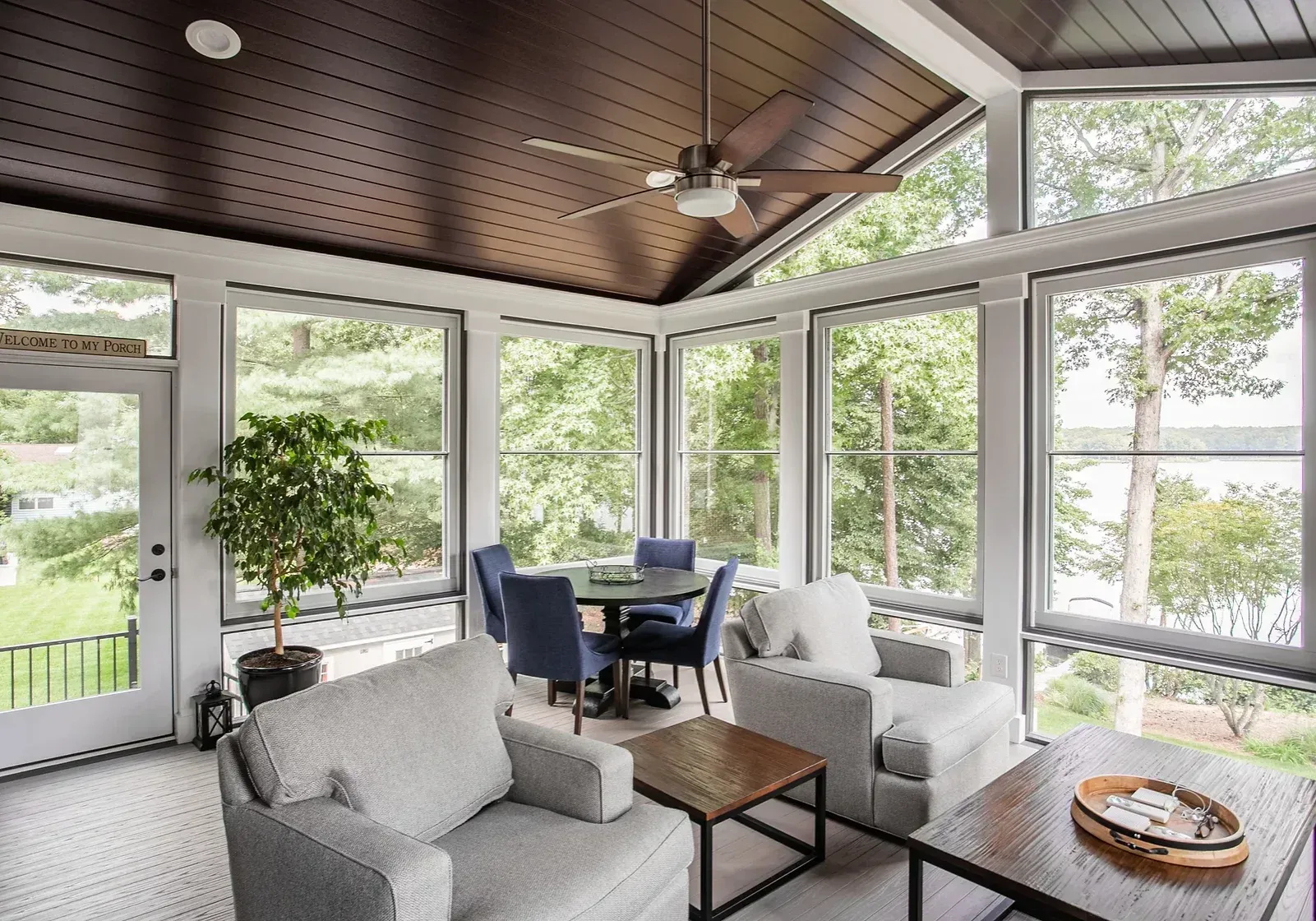 Sunroom with light-colored seating, a table, and a dark wooden ceiling; overlooking a green landscape and lake.