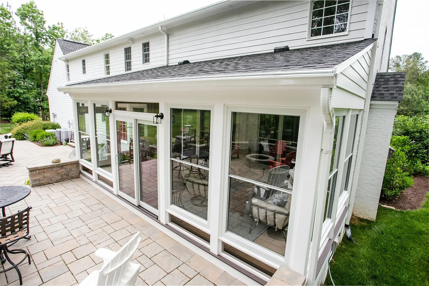 White house with a sunroom featuring large windows, attached to a paved patio, and green lawn.