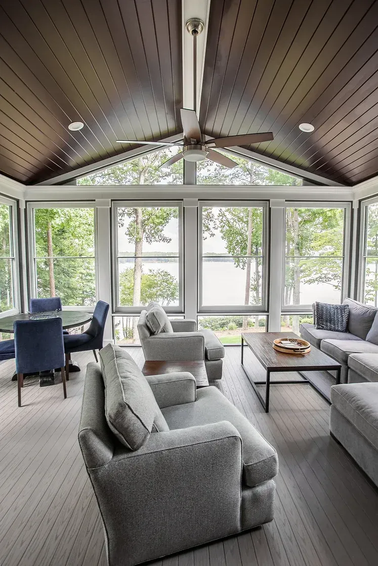 Sunroom with gray sofas, a dining table, and large windows overlooking trees and a lake.