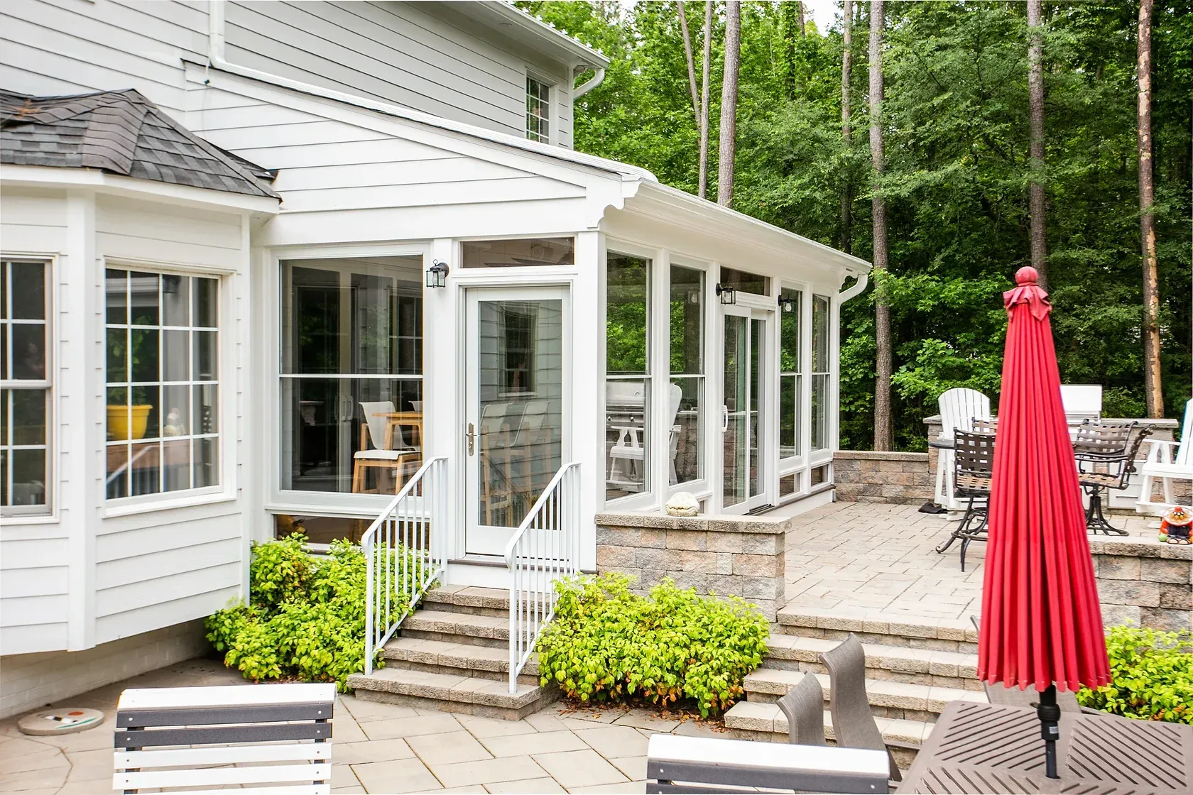 Screened-in porch and patio with white railings, a red umbrella, and greenery, adjacent to a house with trees.