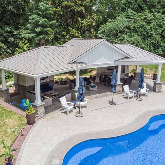 Outdoor patio with pool. White columns, grey roof, seating areas, blue umbrellas, and a curved pool with clear water.