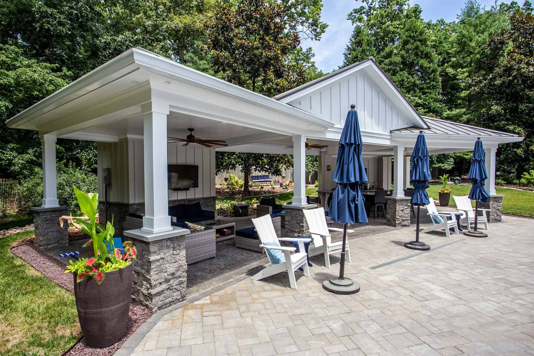 Pavilion with white pillars, stone bases, lounge area, grill, and blue umbrellas on a patio.