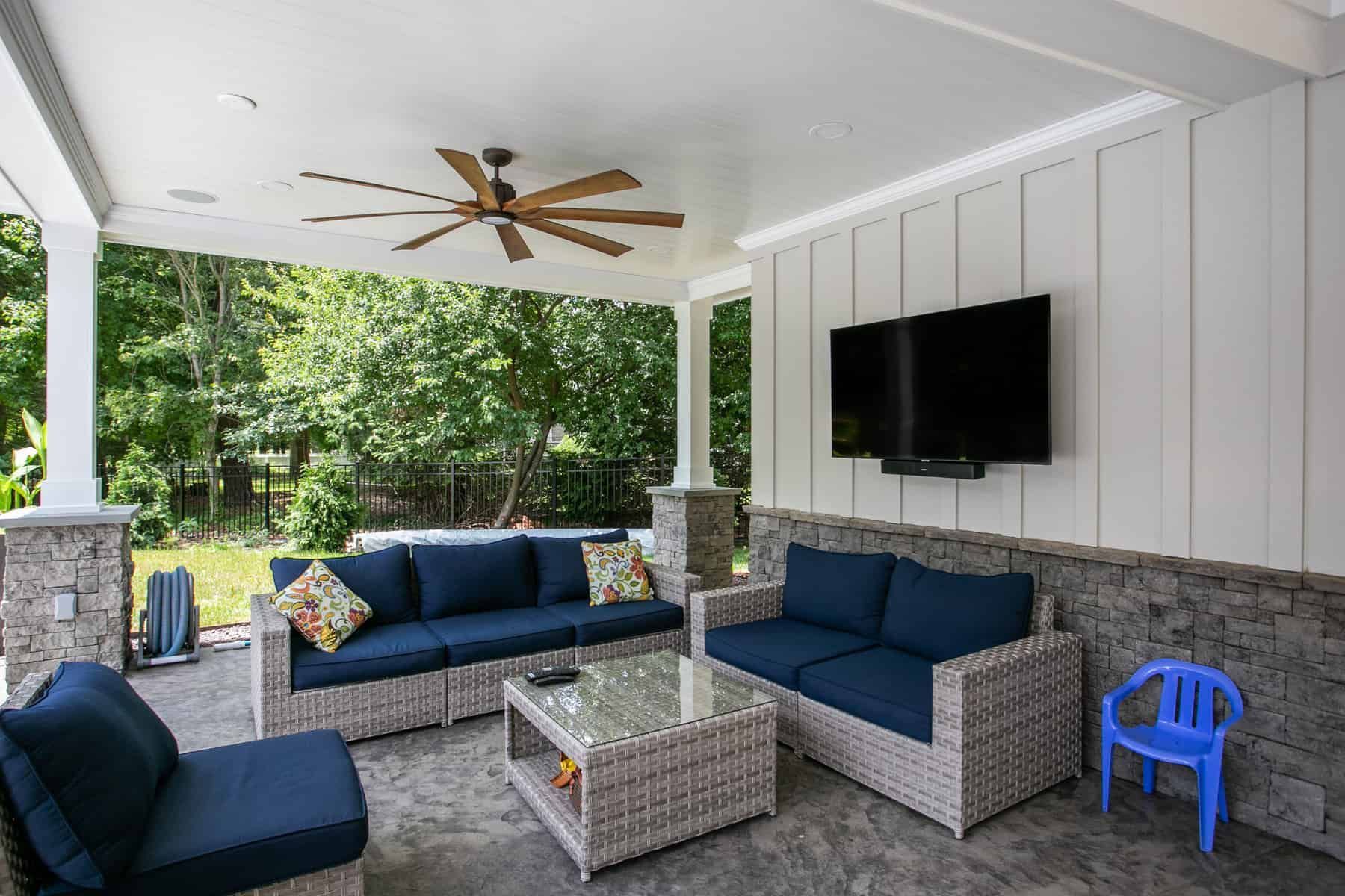 Covered outdoor patio with wicker furniture, TV, ceiling fan, and blue cushions.