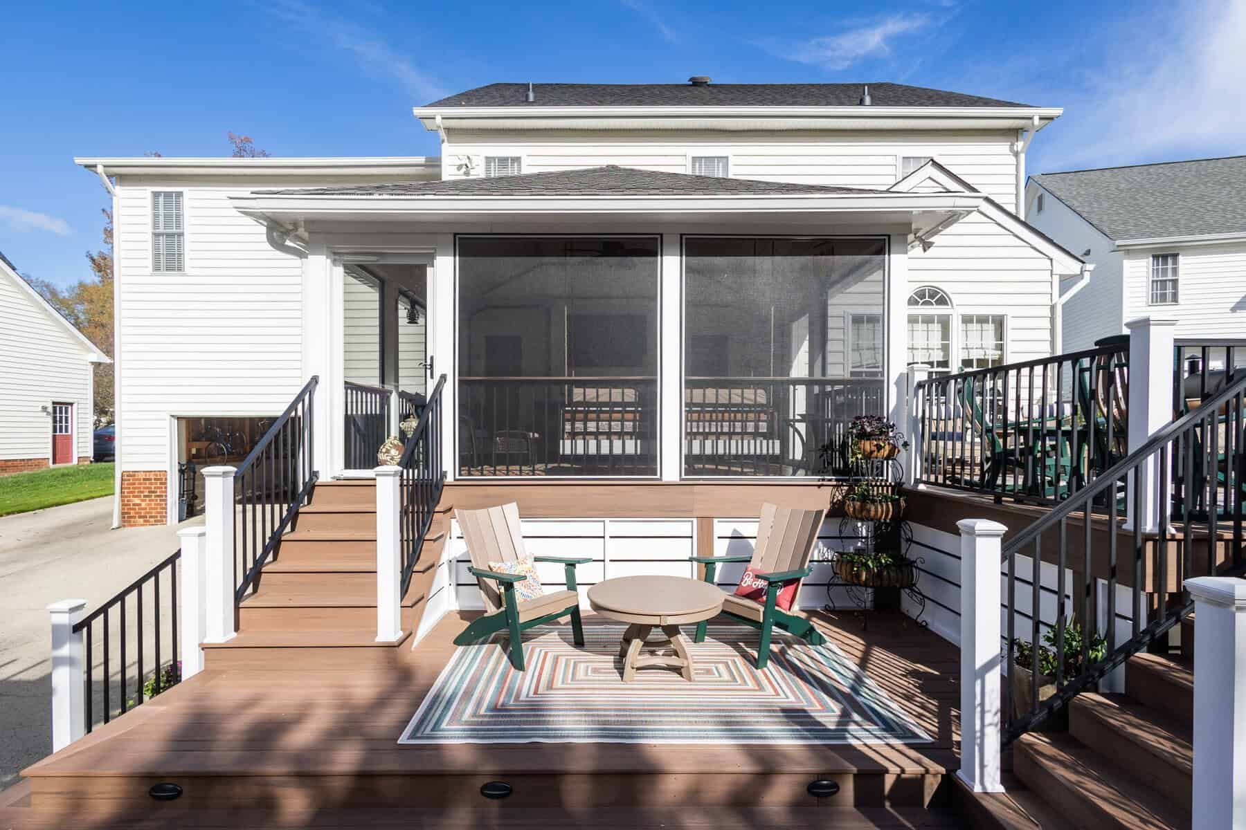 Backyard deck with screened porch, wooden furniture, and black railing against white house.