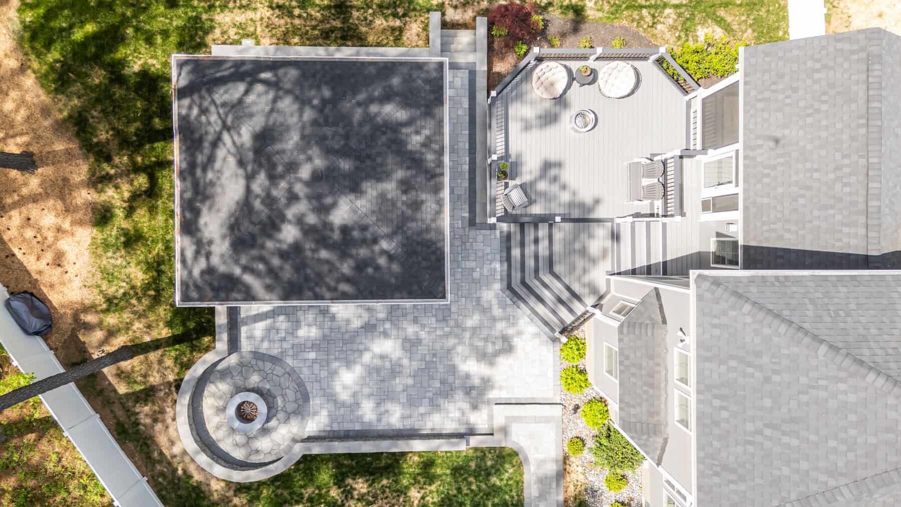 Overhead view of a house with a stone patio, deck, and a fire pit. Gray and brown tones dominate.