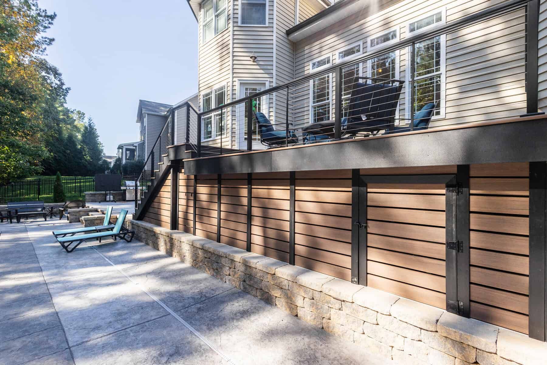 Deck with storage, brown siding, black railing, stone wall, and patio with lounge chairs.