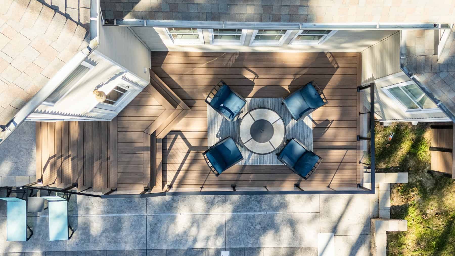 Overhead view of a wooden deck with a central fire pit surrounded by four blue armchairs; adjacent concrete patio.