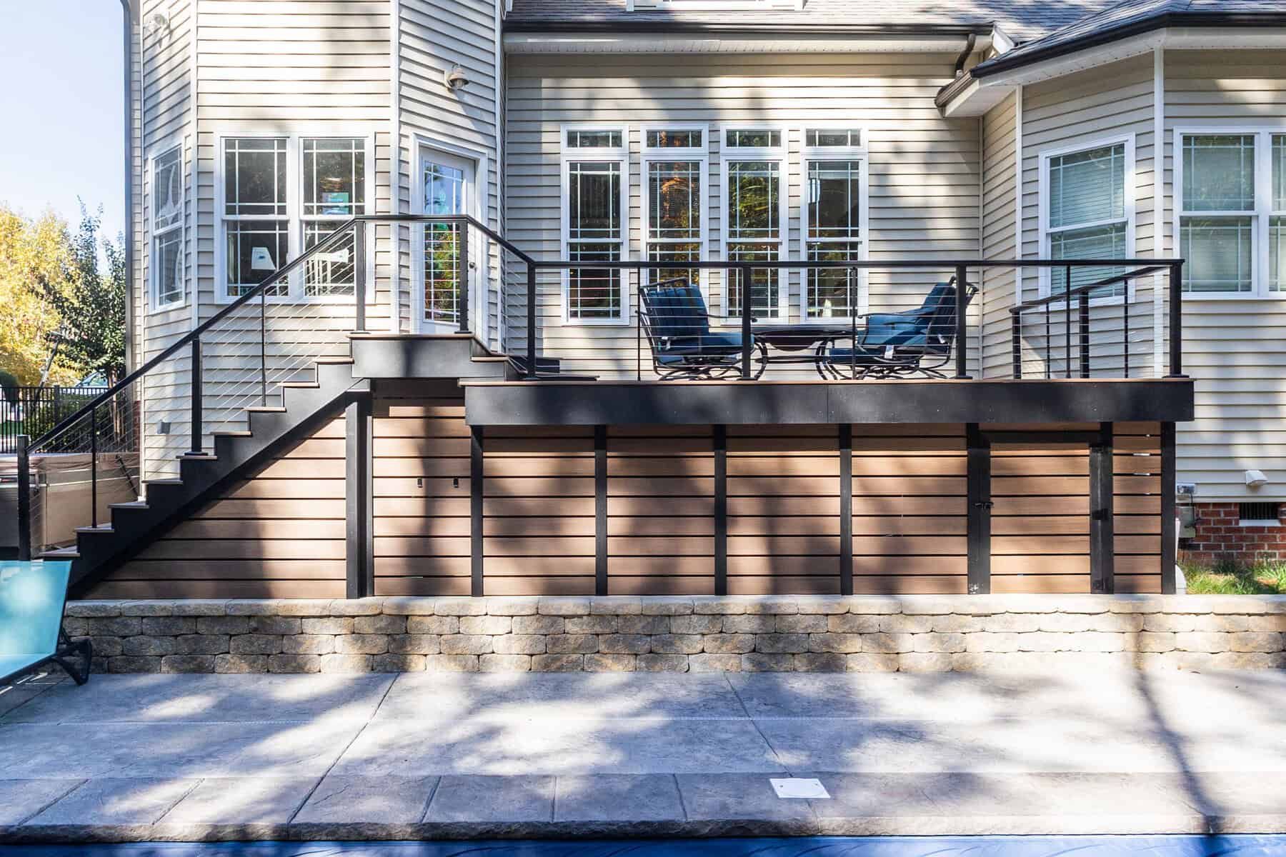 Backyard deck with black railings, tan siding, and horizontal wood panels. Furnished with chairs and table.