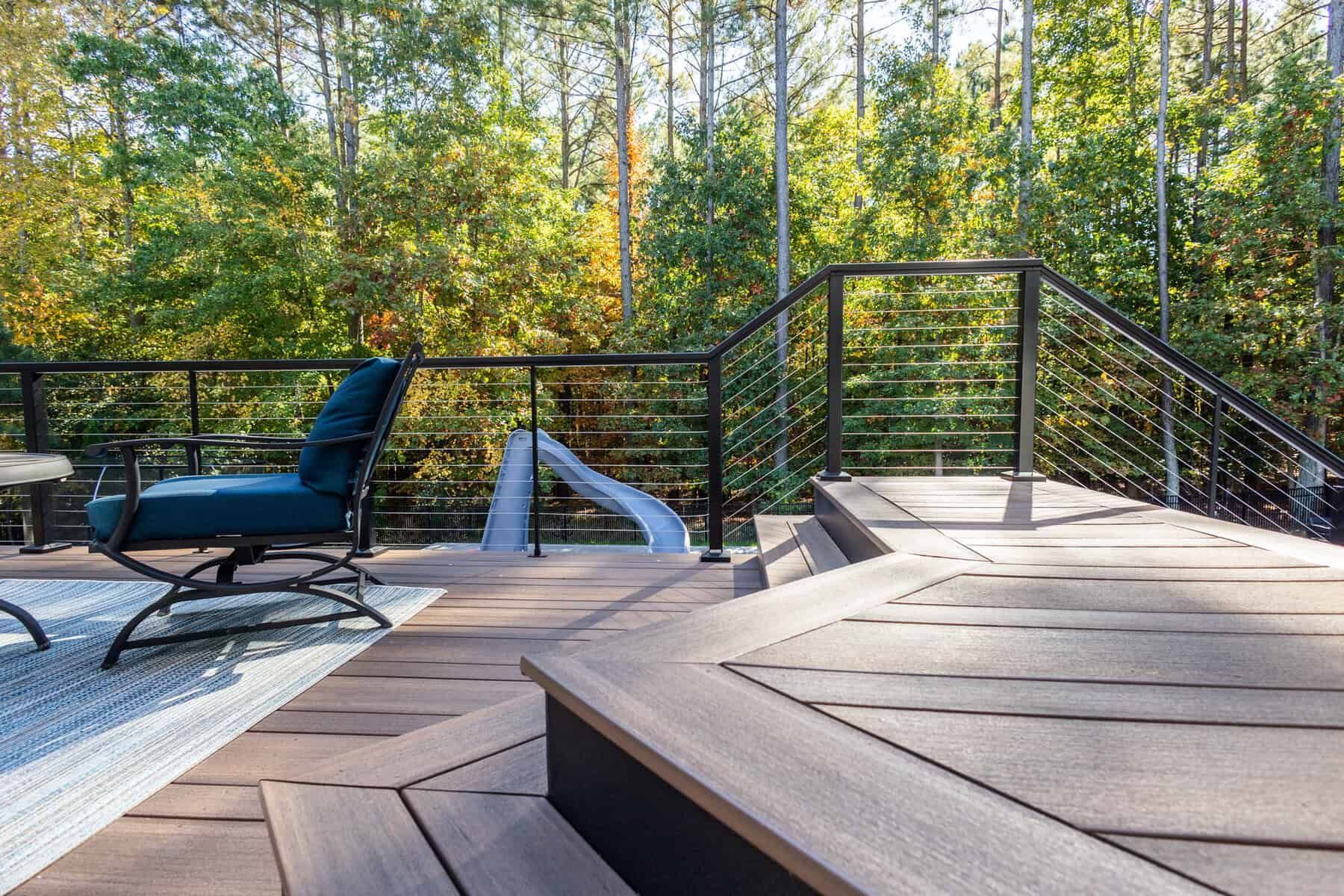 Modern outdoor deck with black railing and steps, blue chair, and trees in background.