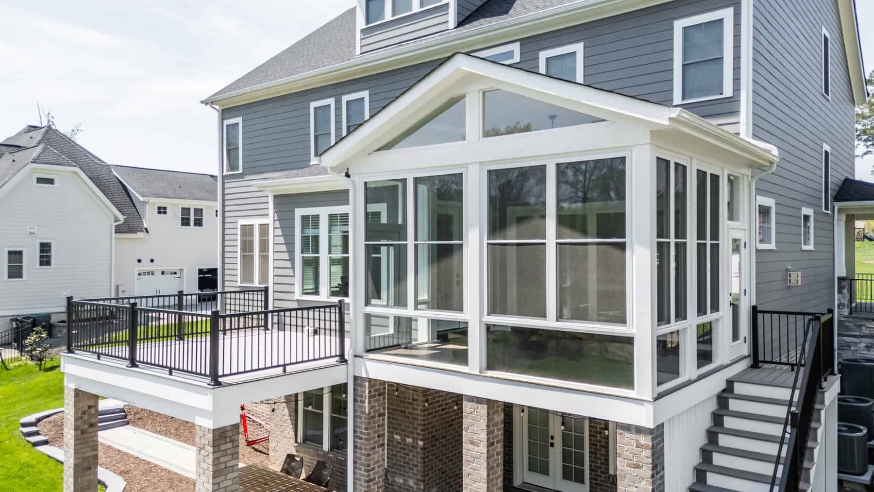 Back of a multi-story house with a deck and enclosed porch; gray siding, brick base.
