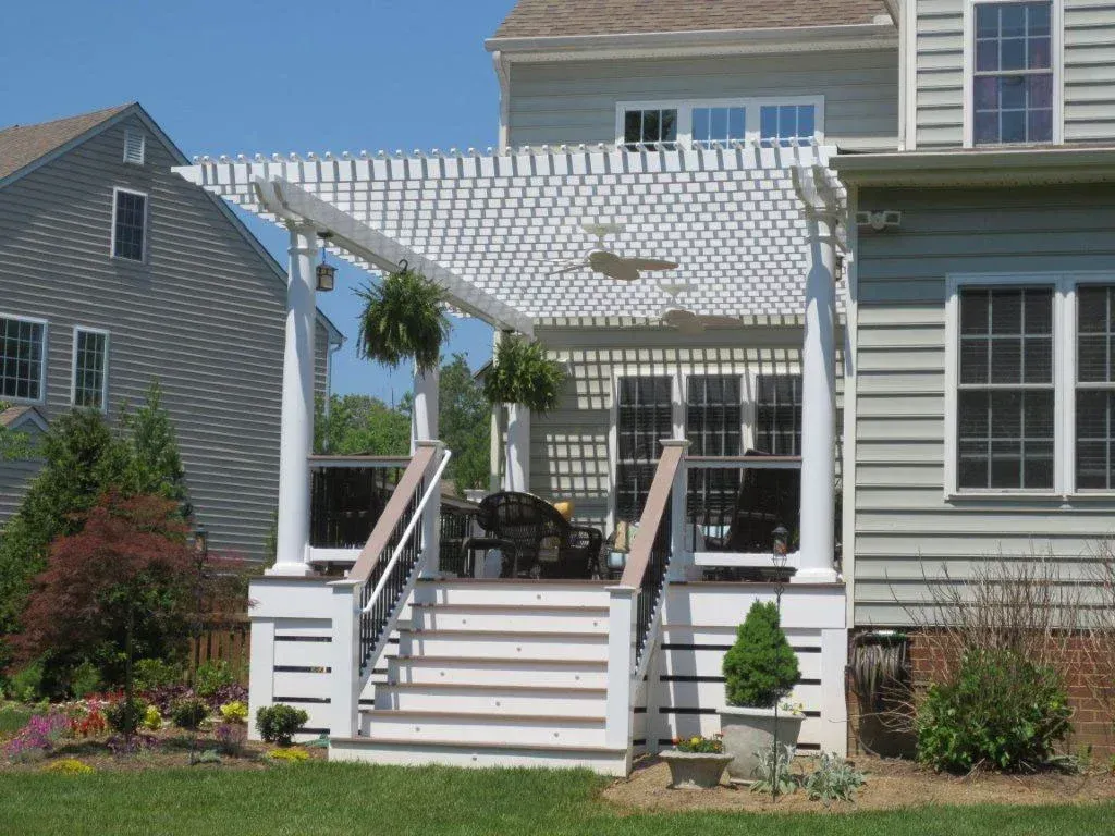 White pergola over a deck with stairs, attached to a light gray house.