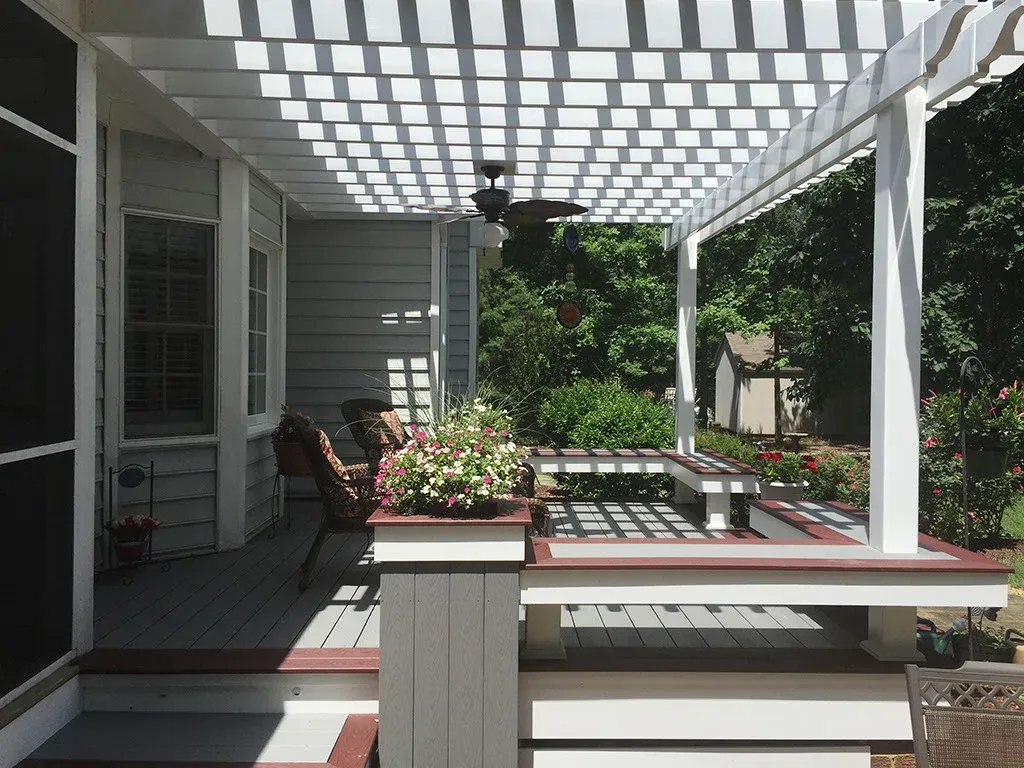 White pergola over a wooden deck with built-in seating, plants, and a ceiling fan.