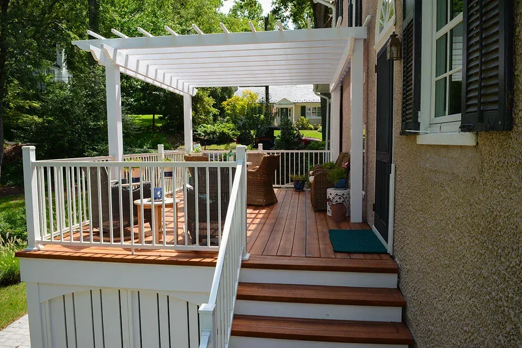 White pergola over a wooden deck with railing and stairs next to a house.