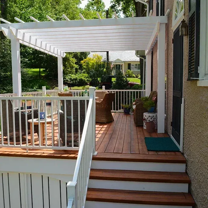 White pergola over a wooden deck with railing and stairs; wicker furniture and green rug are present.