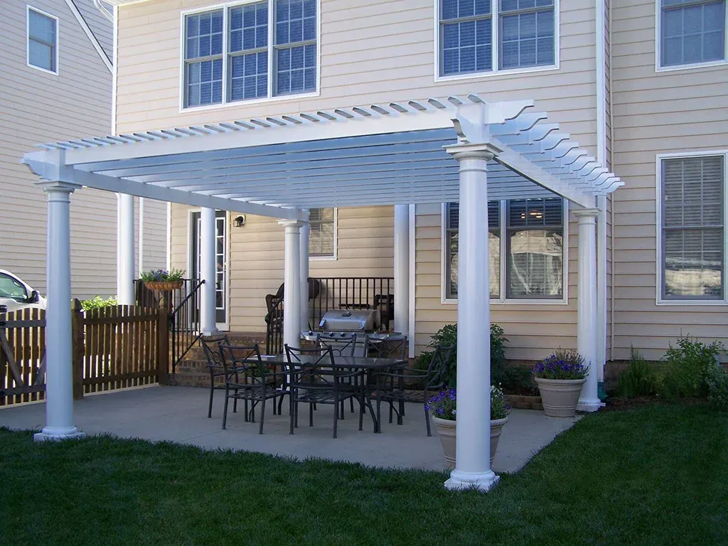 Pergola over patio with table and chairs, adjacent to a house with windows.