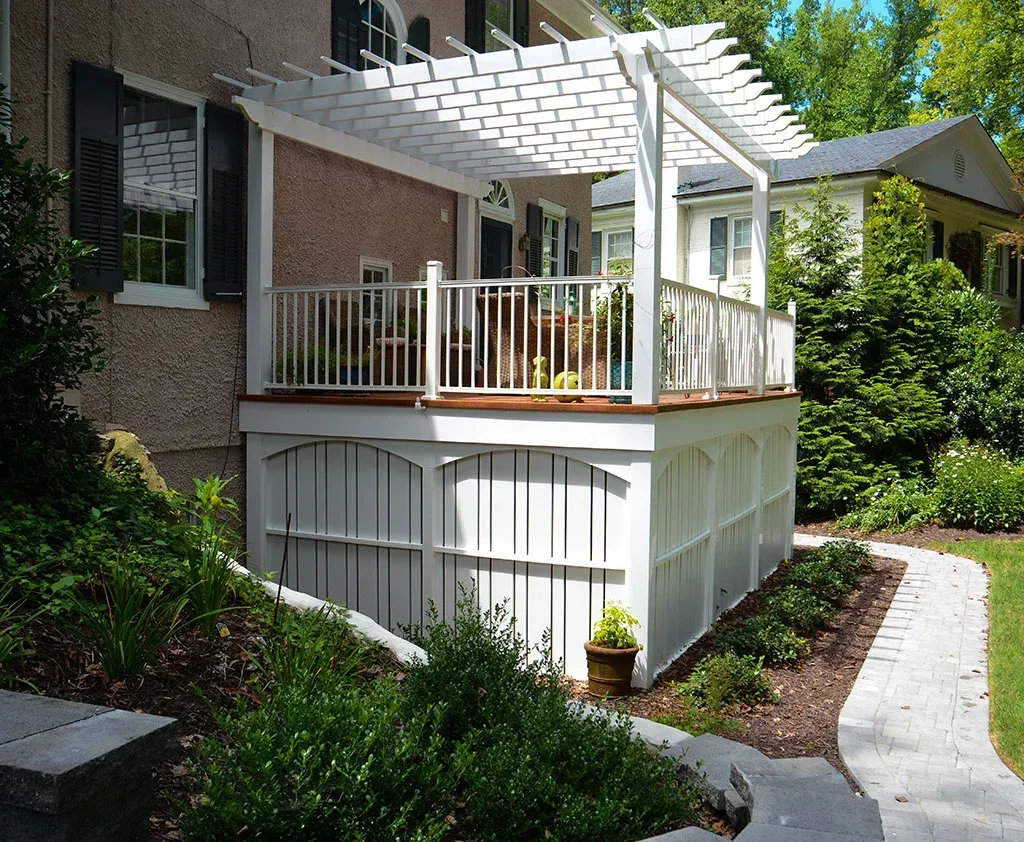 White pergola and deck with railing attached to a house. Landscaping in the foreground.