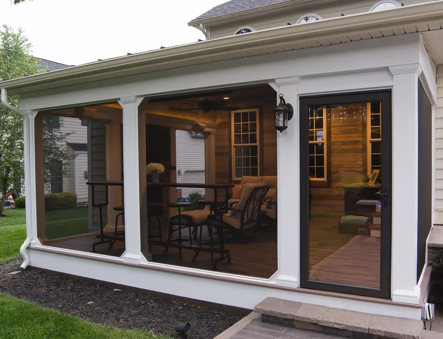 Screened-in porch with white columns, brown screen, and door leading inside. Interior features furniture and lights.