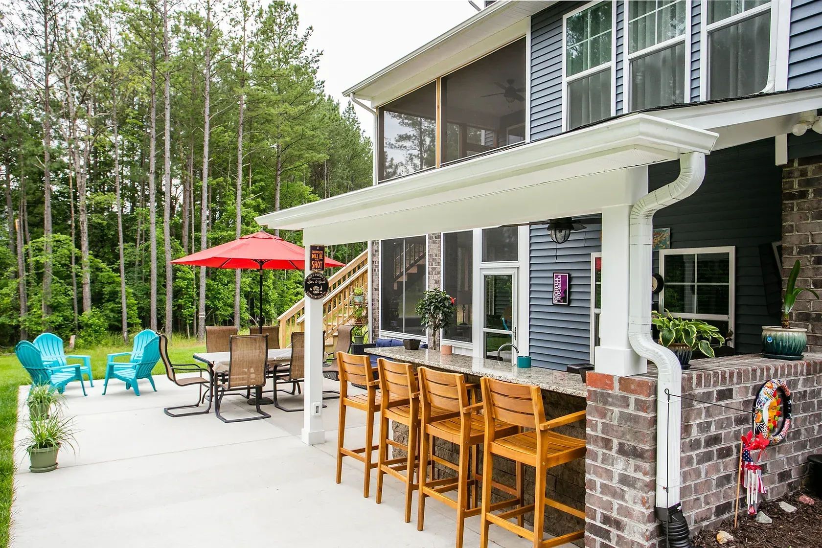 Outdoor patio with a bar, seating, and trees in the background. Blue house with a screened porch is adjacent.