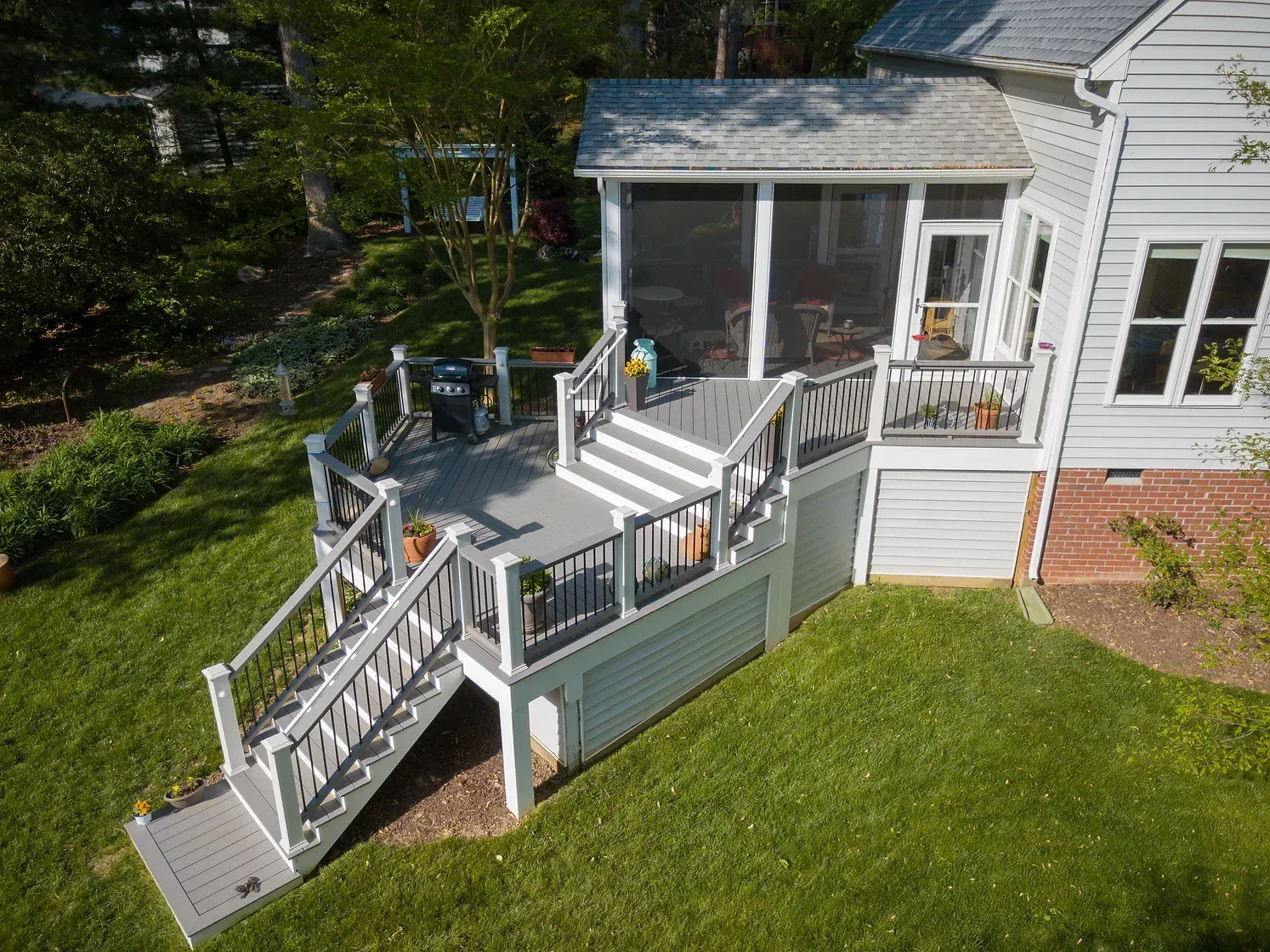 Grey and white tiered deck with stairs, a screened porch, and a grill, built on a grassy slope.