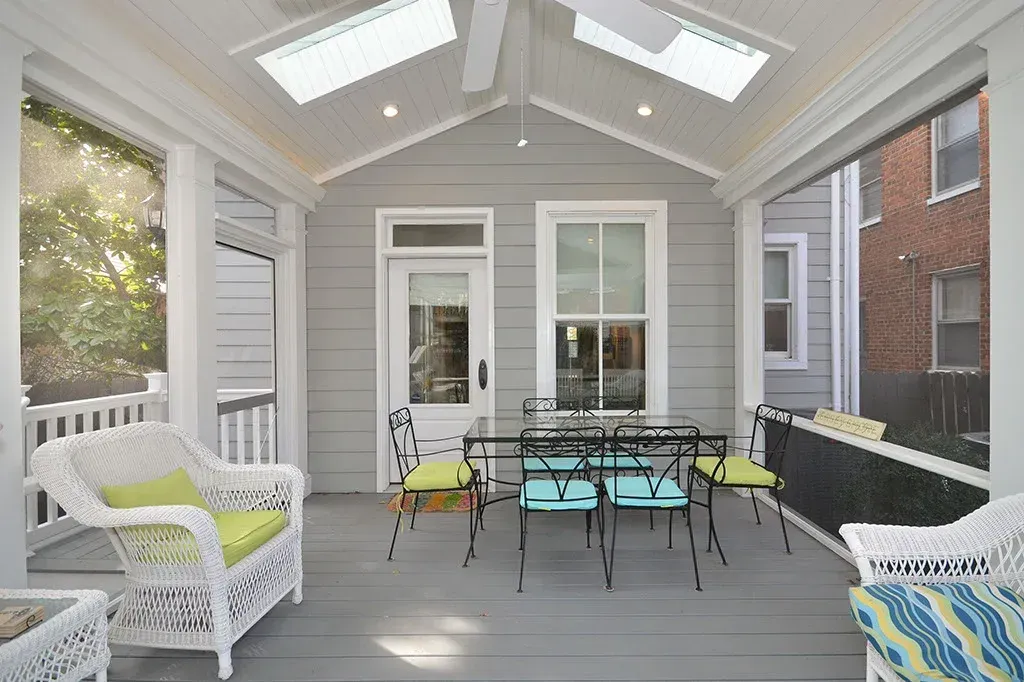 Screened-in porch with white wicker furniture, dining table with colorful chairs, and skylights; gray walls and floor.
