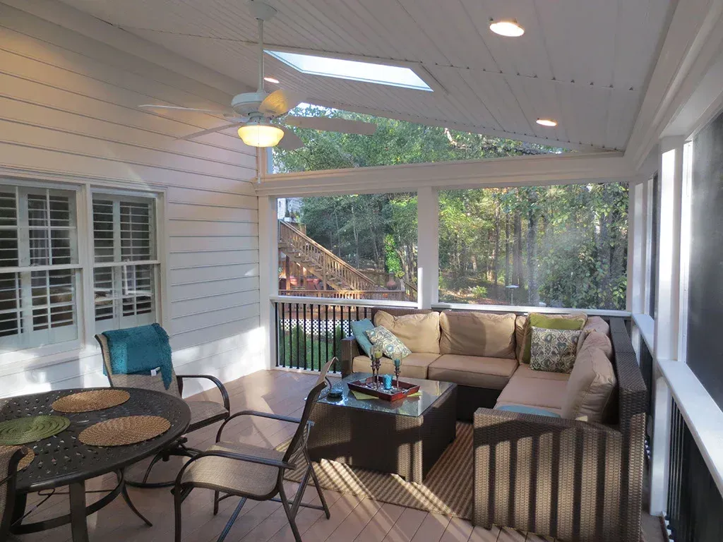 Screened-in porch with sectional sofa, table, chairs. Sunlight streams through the skylight, trees visible through the screen