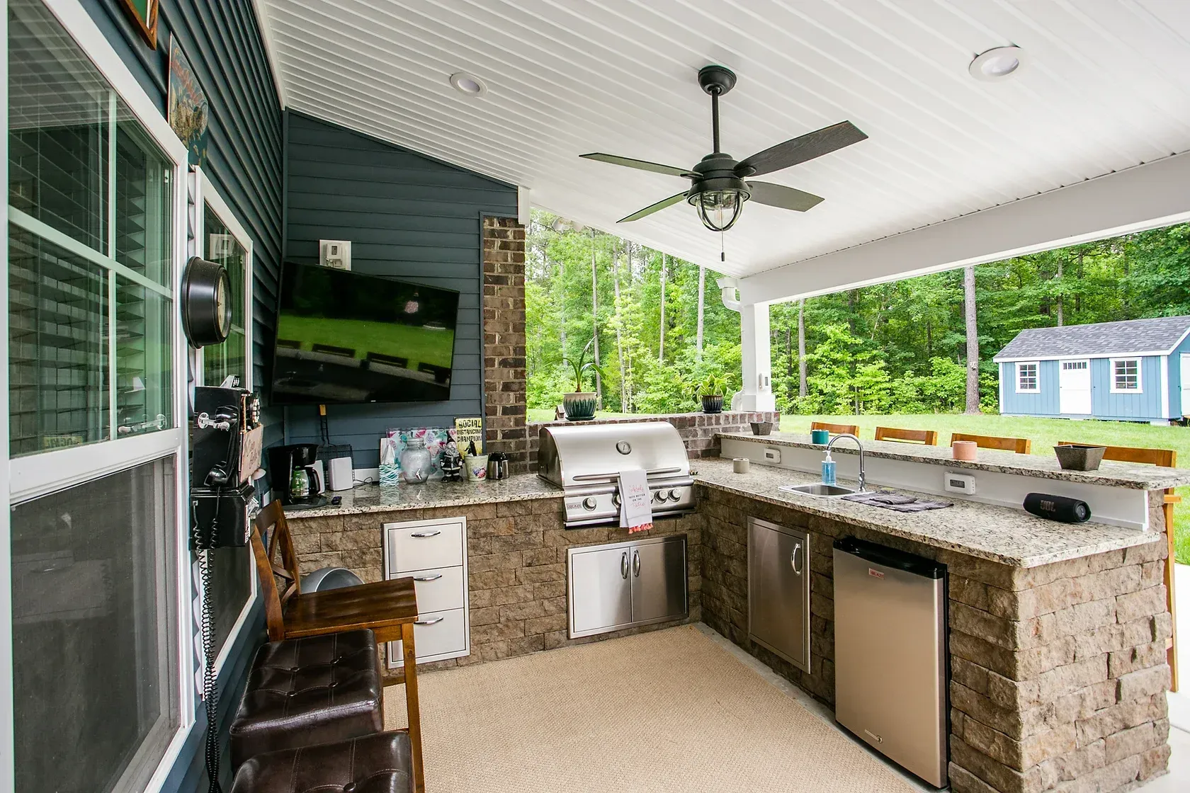 Outdoor kitchen with a grill, sink, refrigerator, and TV; surrounded by a deck and trees.
