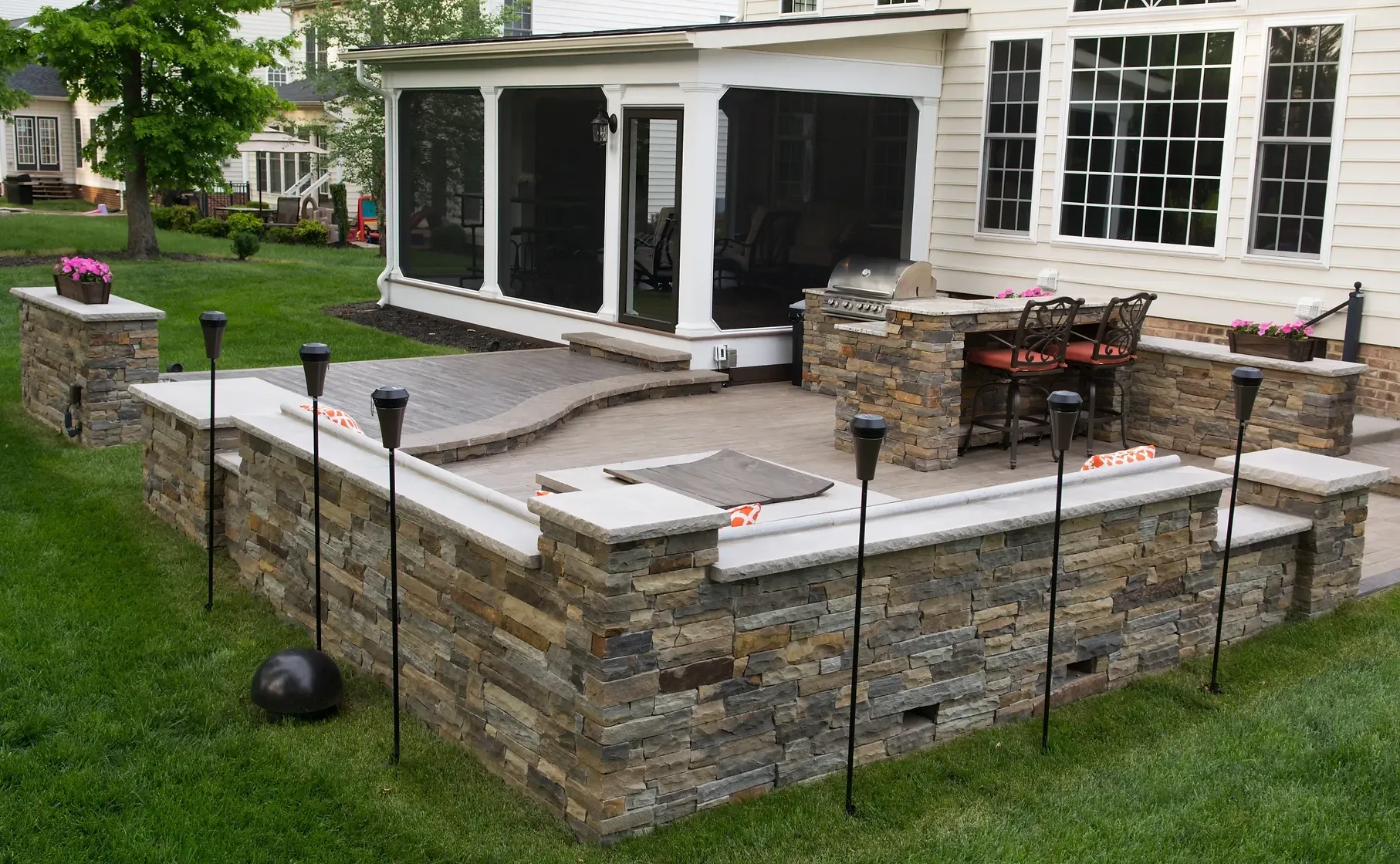 Stone patio with built-in grill and seating, next to a screened porch, surrounded by a low wall with tiki torches.
