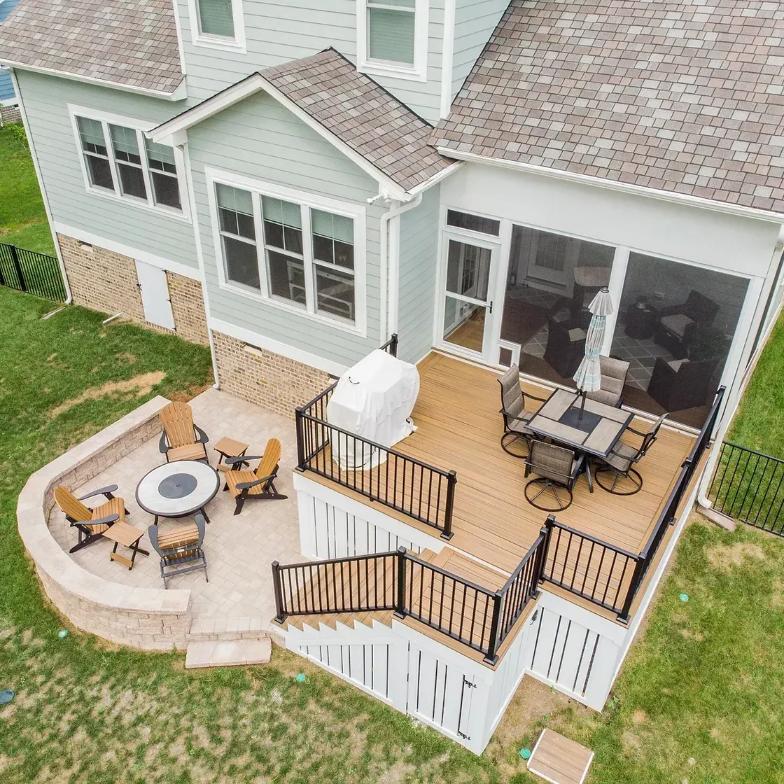 Aerial view of a home with a deck and patio. Deck has a dining set. Patio features a fire pit with seating.