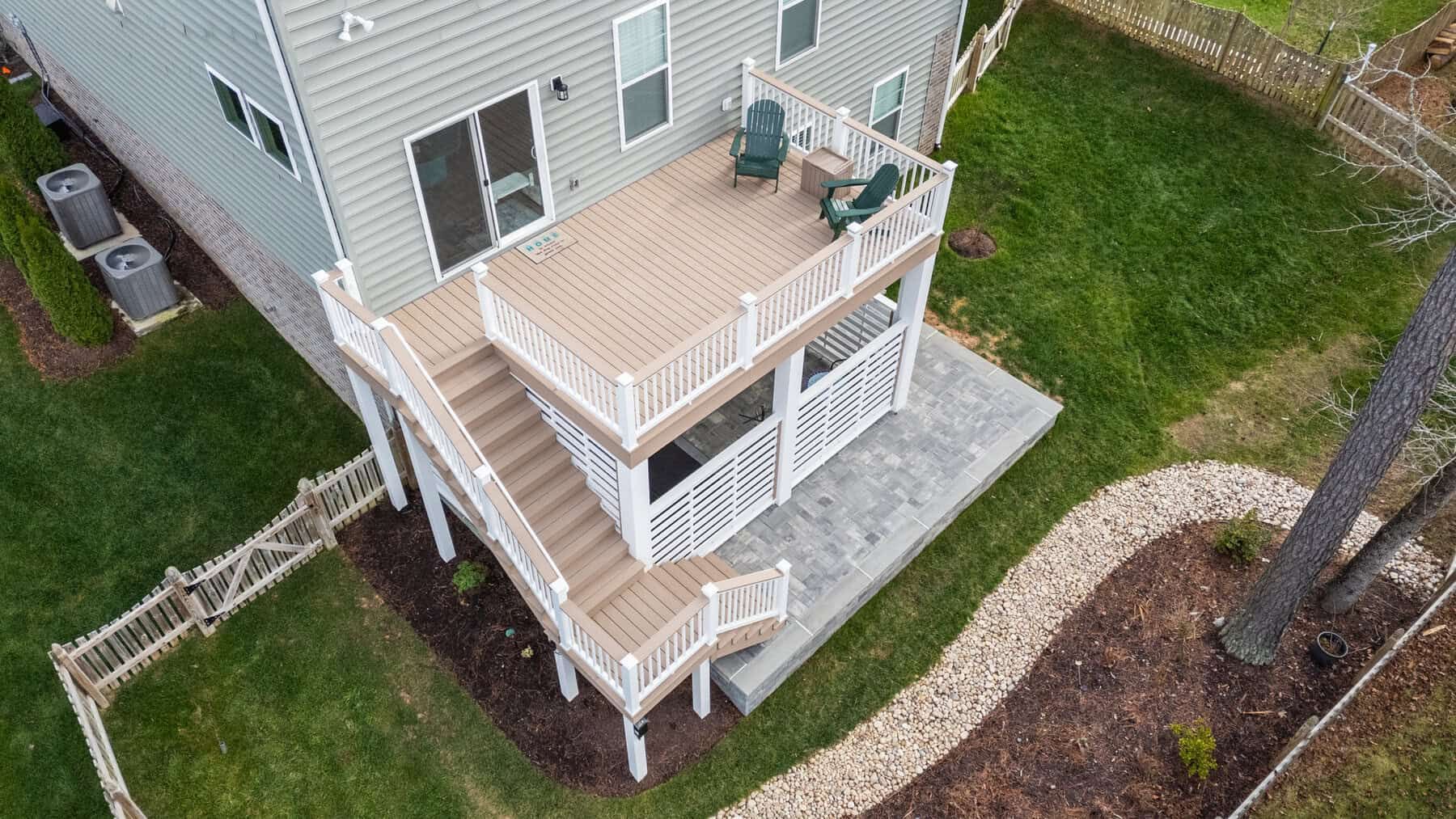 Two-level deck with stairs, built against a gray house. White railing, tan decking, green lawn, and patio below.