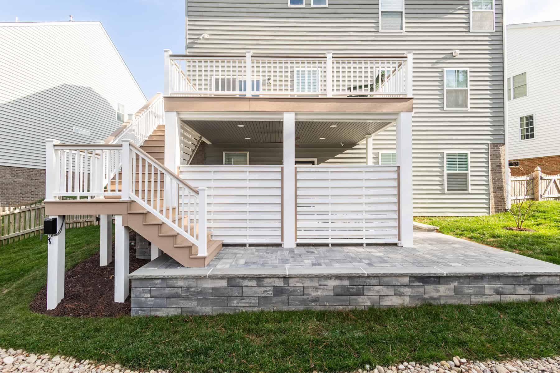 Rear view of a two-story deck with stairs, a patio, and a light gray house.
