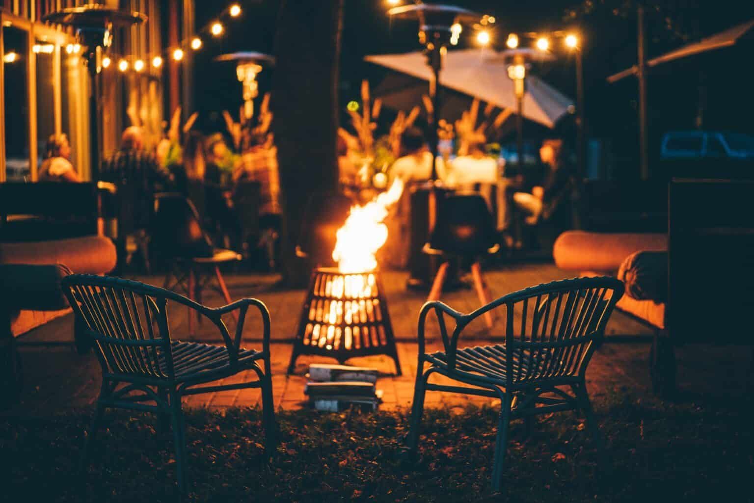 Two wicker chairs face a bonfire in a dimly lit outdoor setting with string lights and heaters.