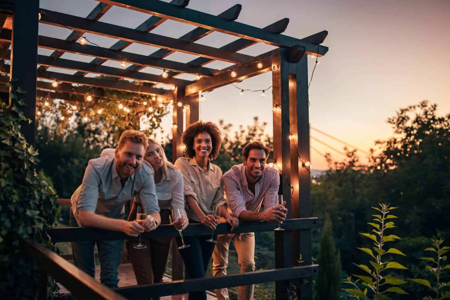 Four people smile, leaning on a wooden deck, strings of lights overhead, sunset in background.