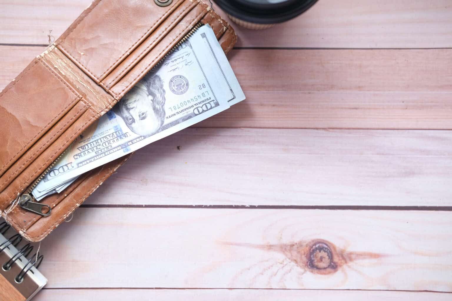 Brown wallet open, displaying several one hundred dollar bills on wooden surface.