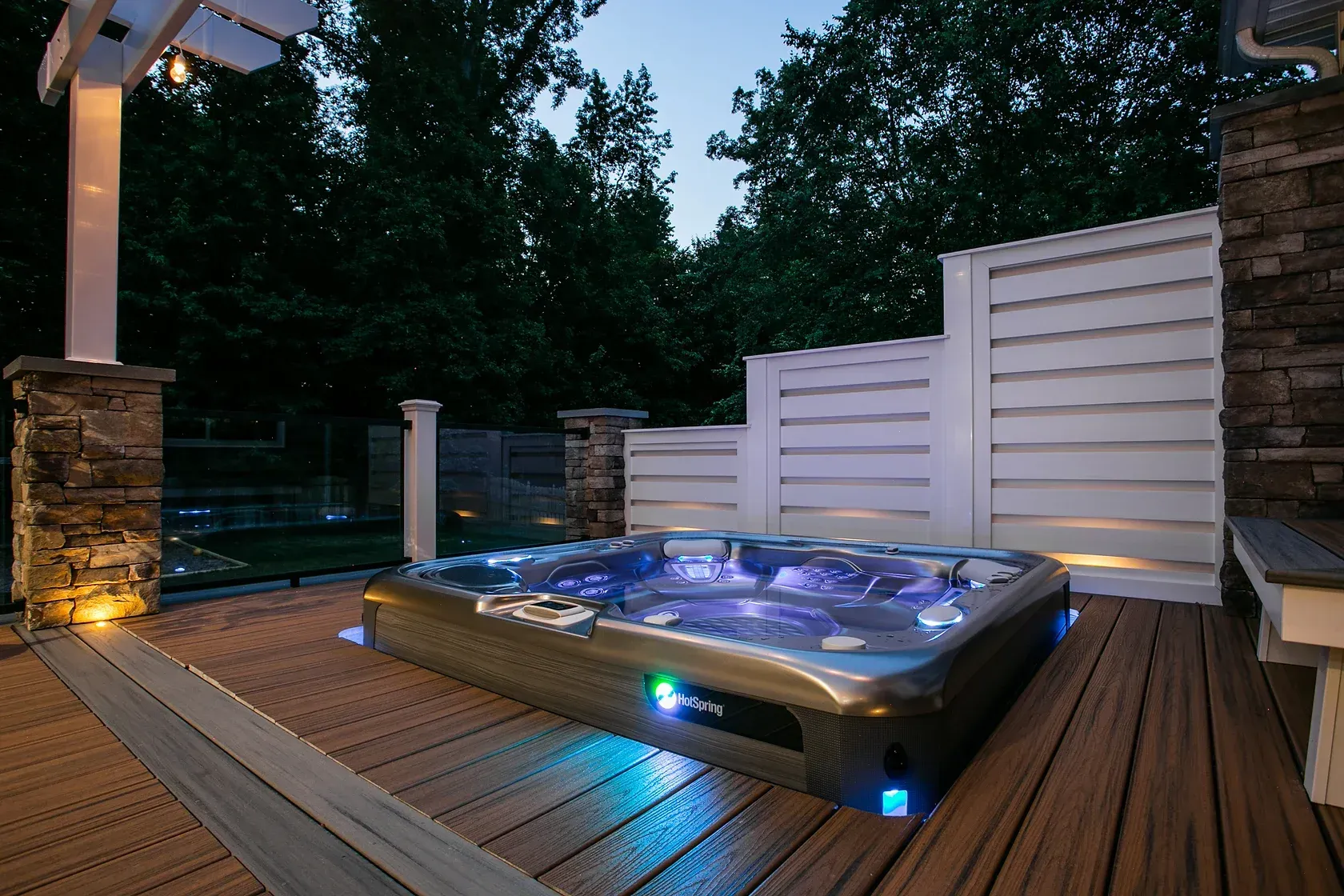 A lit hot tub on a wooden deck at dusk, with white fence and trees in background.