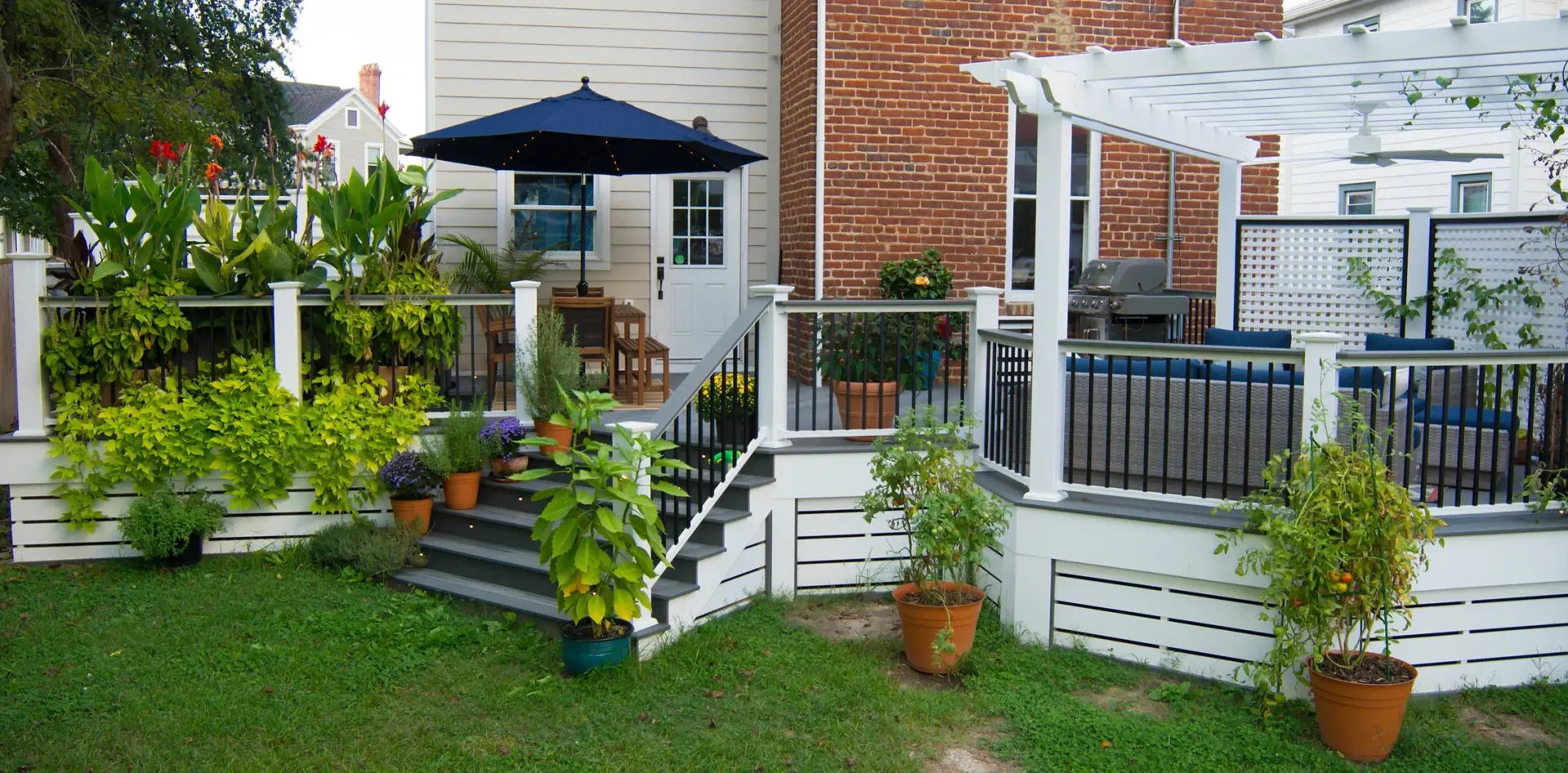 A backyard patio with a deck, plants, stairs, and a dark blue umbrella.