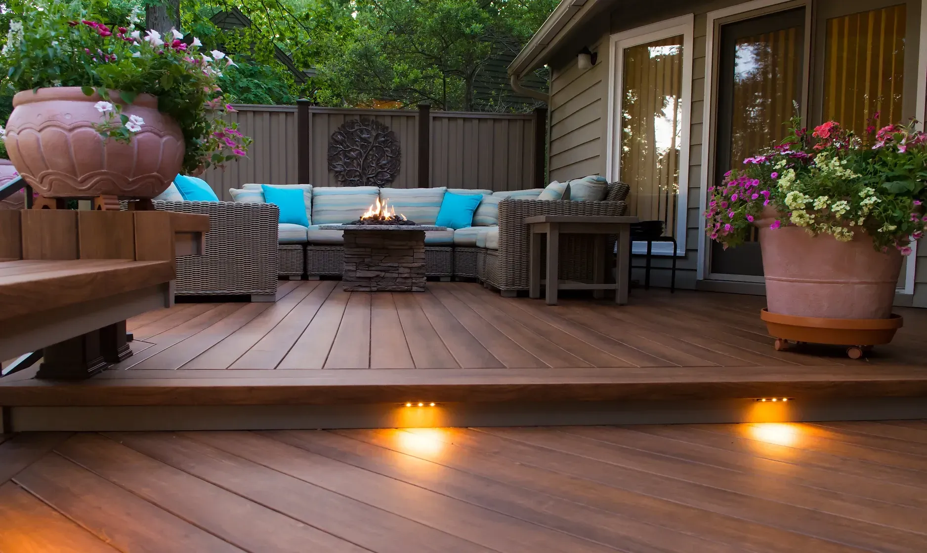 Wooden deck with outdoor seating, a fire pit, and potted plants lit by warm spotlights.
