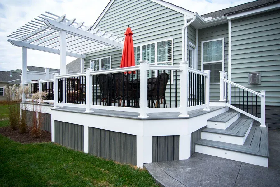 White deck with black railings, pergola, umbrella, and steps next to a house with gray siding.