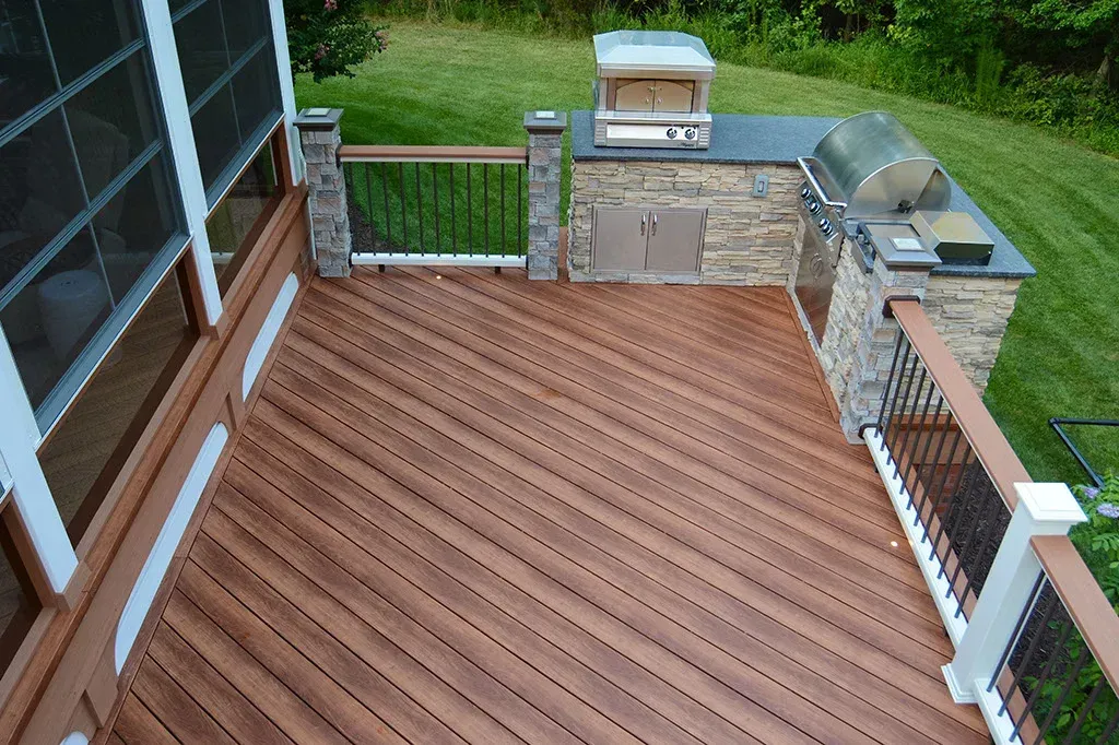 Wooden deck with built-in outdoor kitchen, including grill and pizza oven, next to a screened porch.