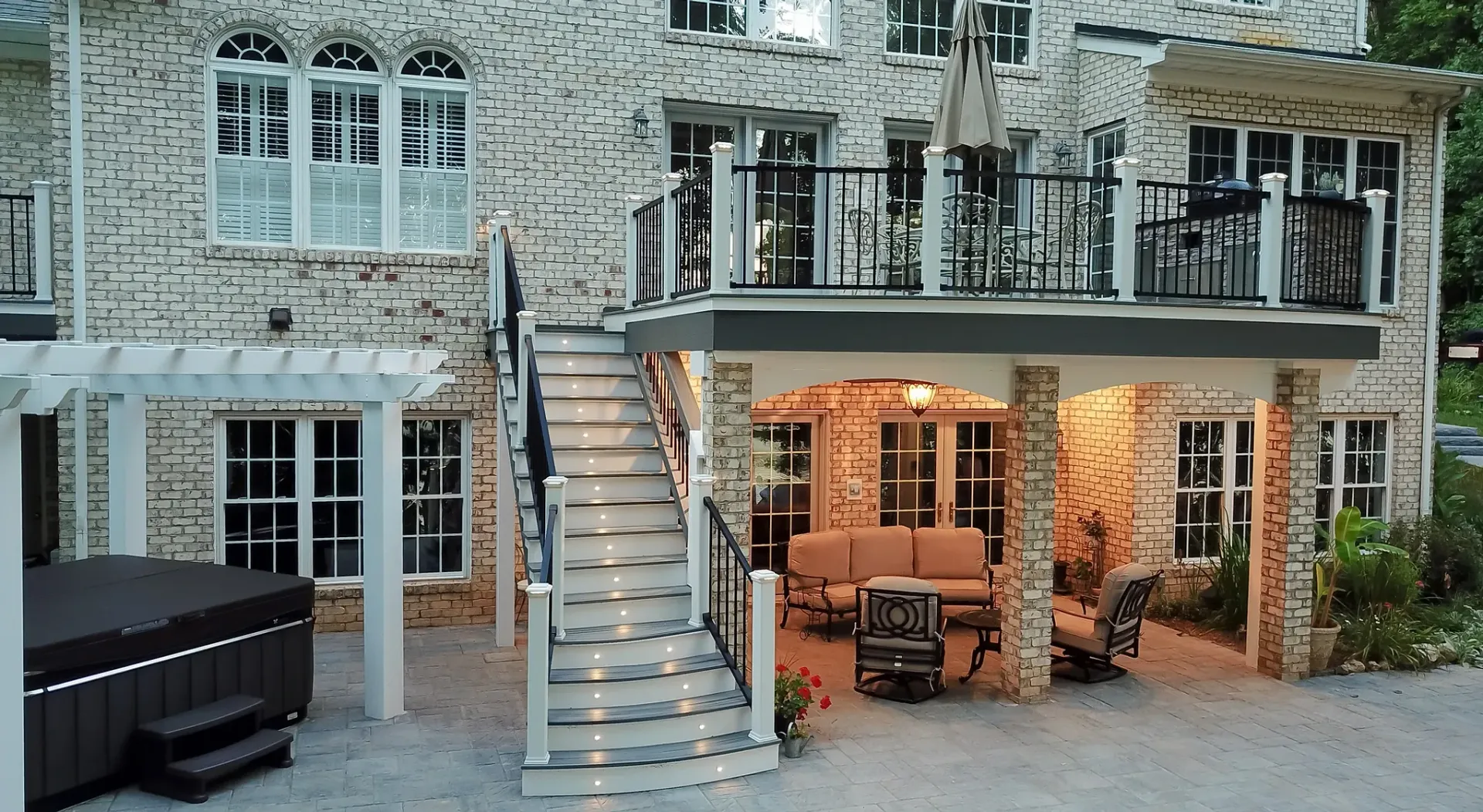 Backyard with multi-level deck, hot tub, and stone patio under elevated balcony, beige brick exterior.
