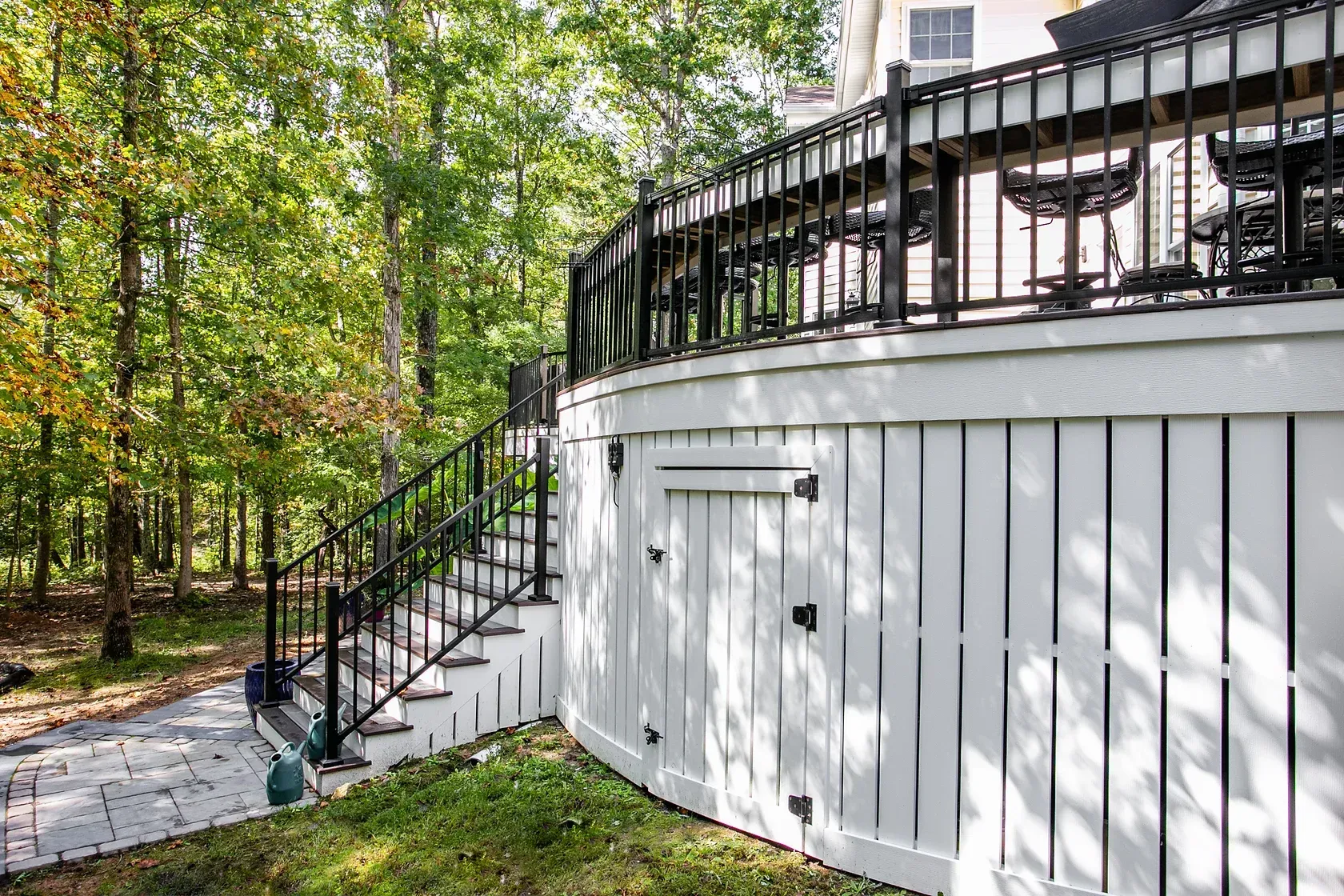 White-sided deck with black railings and steps leading to a stone path, surrounded by trees.