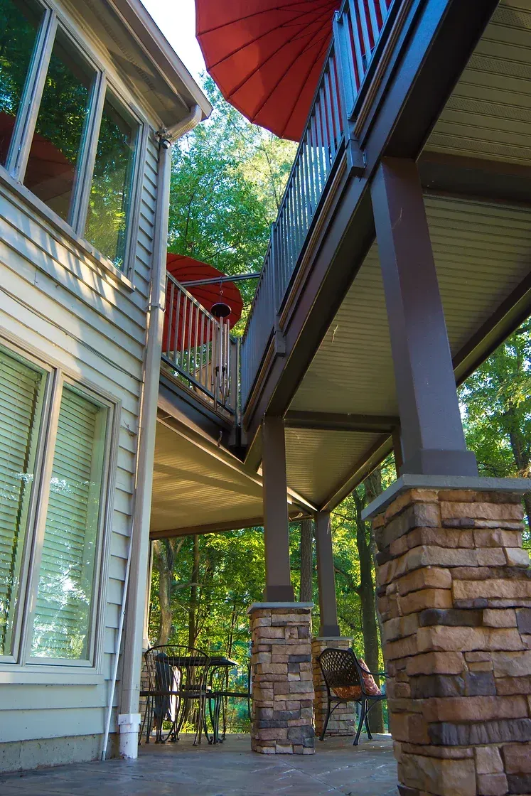 Exterior view of a house with a raised deck supported by stone columns; red umbrellas shade the upper deck.