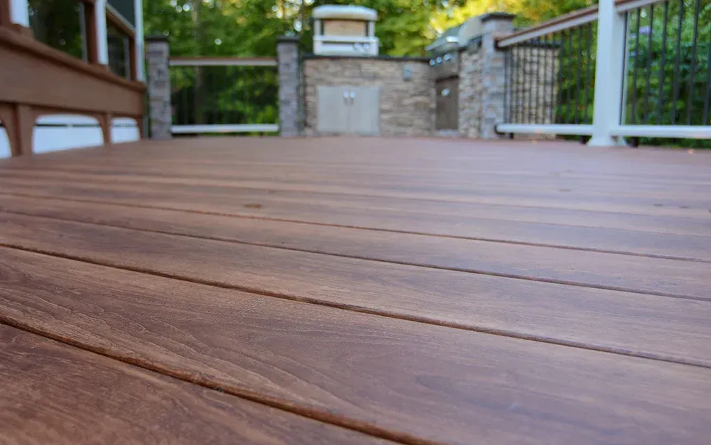 Close-up view of a brown composite deck with a blurred outdoor kitchen and railing in the background.