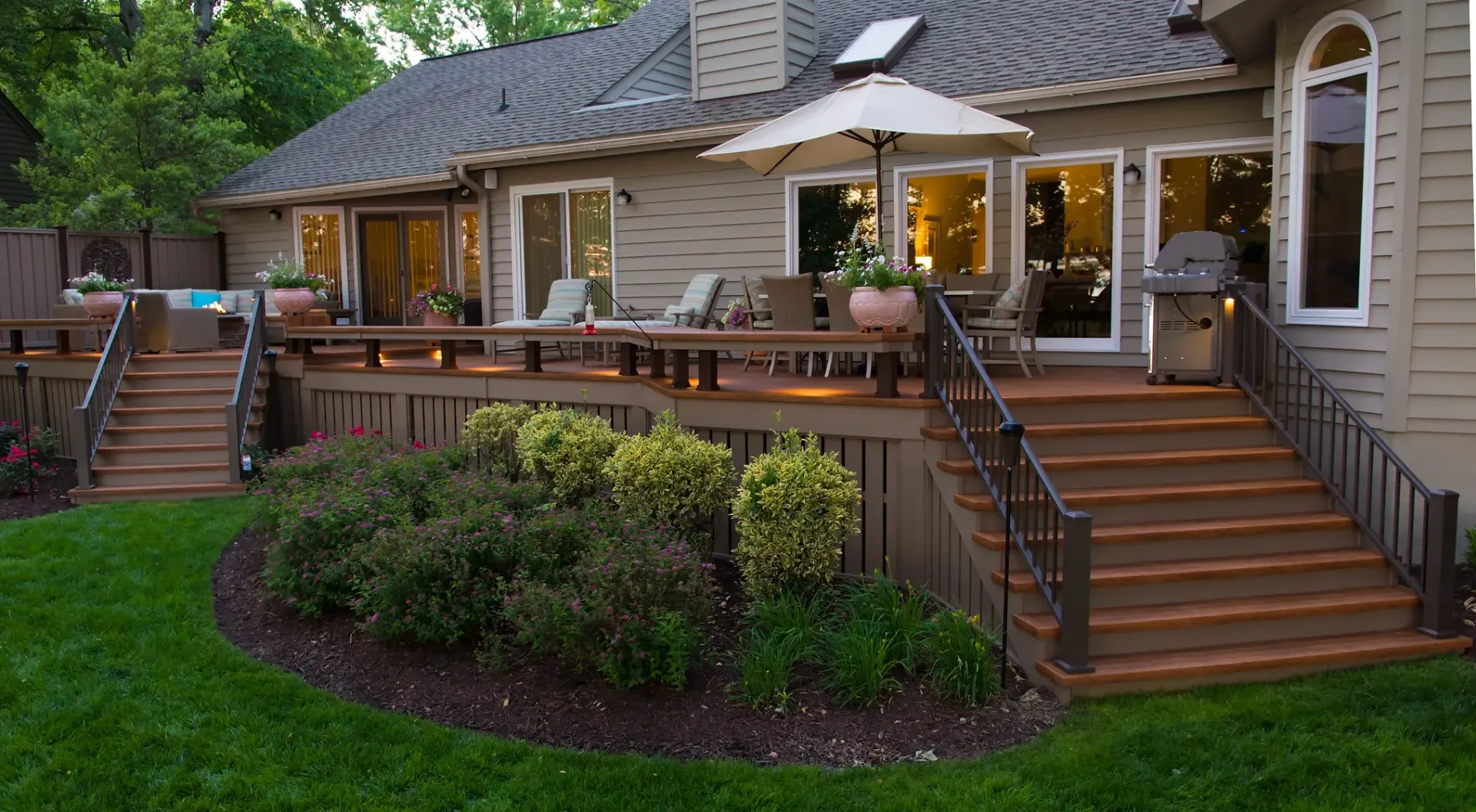 A backyard deck with stairs, surrounded by a lawn and landscaping, beside a house with glass doors.