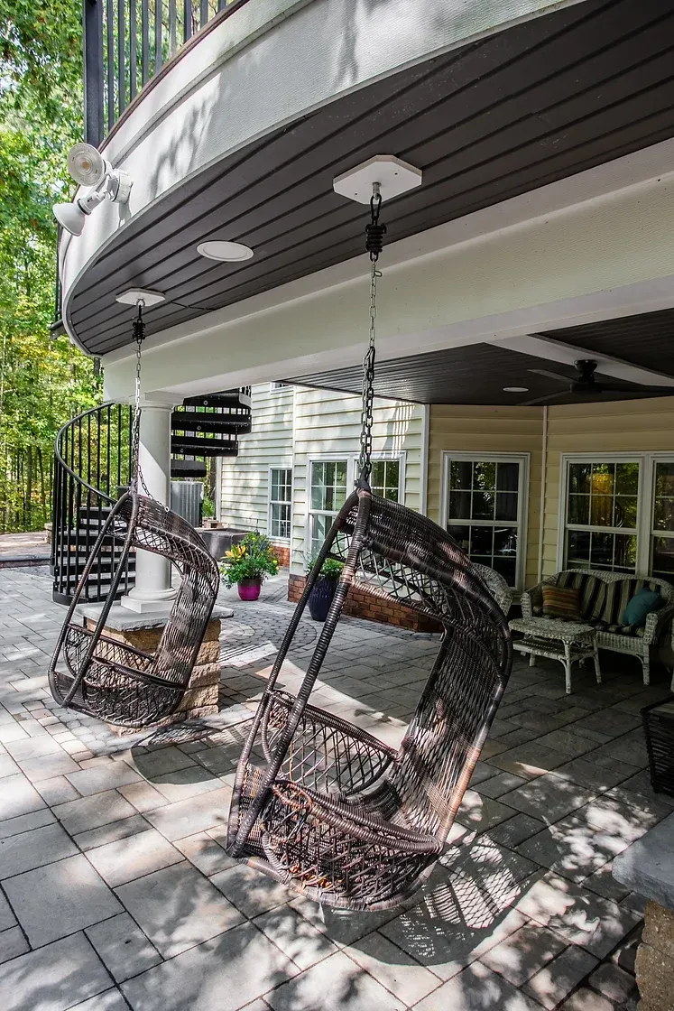 Two hanging wicker chairs on a patio under a curved awning, near a spiral staircase and a house.
